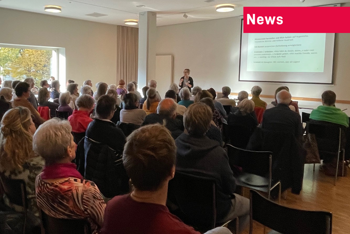 Zahlreiche Menschen hören einem Referat zu. Aufnahme von ganz hinten im Saal. Man sieht auch die Referentin und die Leinwand mit einer Präsentation