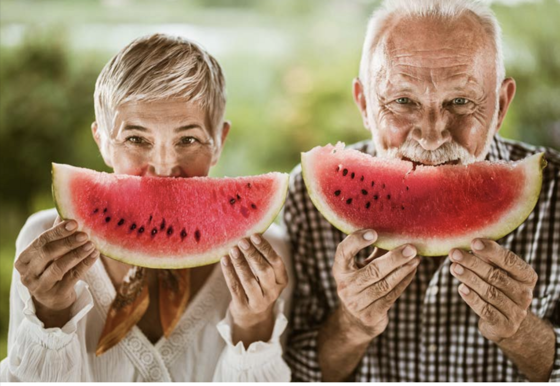 Zwei Personen, Frau und Mann mit grauen oder weissen Haaren, beide halten sich einen Wassermelonen-Schnitz vors Gesicht, so dass der Wassermelonen-Schnitz ein wenig wie ein Lächeln ausschaut.