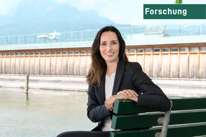 Ein Portrait von Karin Stadelmann, Sie sitzt auf einer Parkbank am See und trägt einen eleganten Blazer. Im Hintergrund eine berühmte Luzerner Brücke