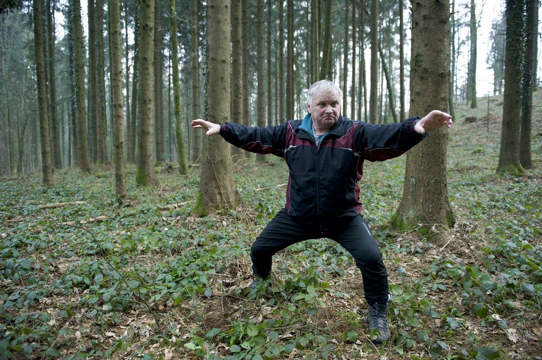 Ein älterer Herr im altmodischen, zweiteiligen Trainingsanzug steht mitten im Wald und macht eine Karate-Pose