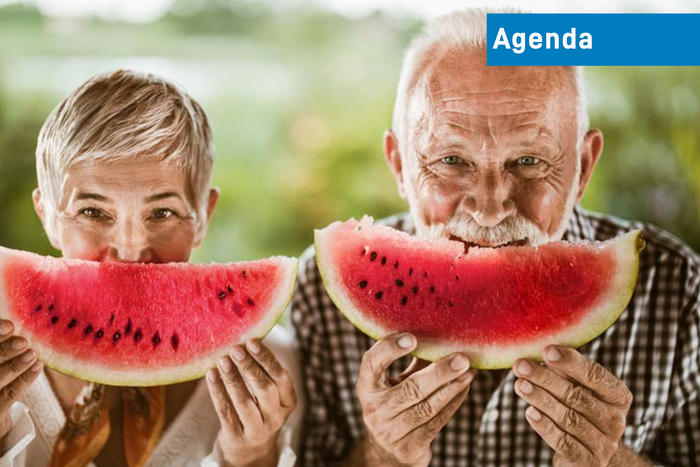 Zwei Personen, Frau und Mann mit grauen oder weissen Haaren, beide halten sich einen Wassermelonen-Schnitz vors Gesicht, so dass der Wassermelonen-Schnitz ein wenig wie ein Lächeln ausschaut.